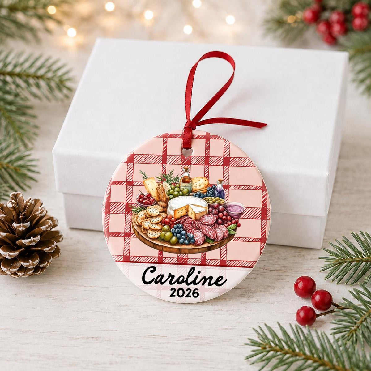 Personalized charcuterie board ceramic ornament with name and year on red plaid in front of a white gift box with pinecones and red berries