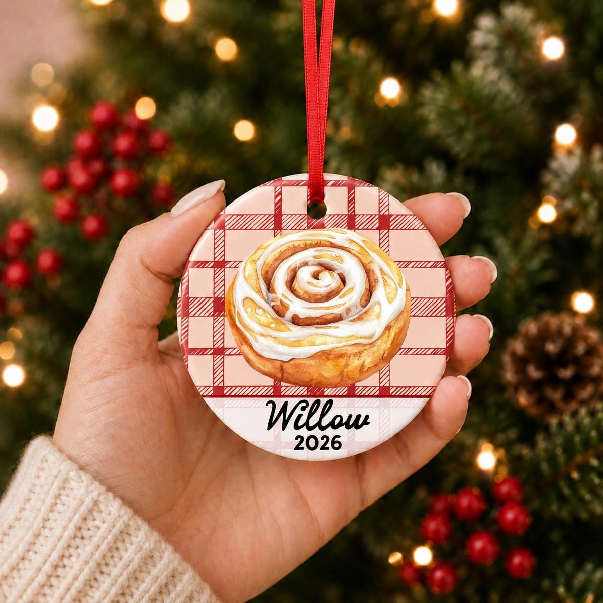 Hand holding the personalized cinnamon roll ceramic ornament with name and year on red plaid in front of a Christmas tree