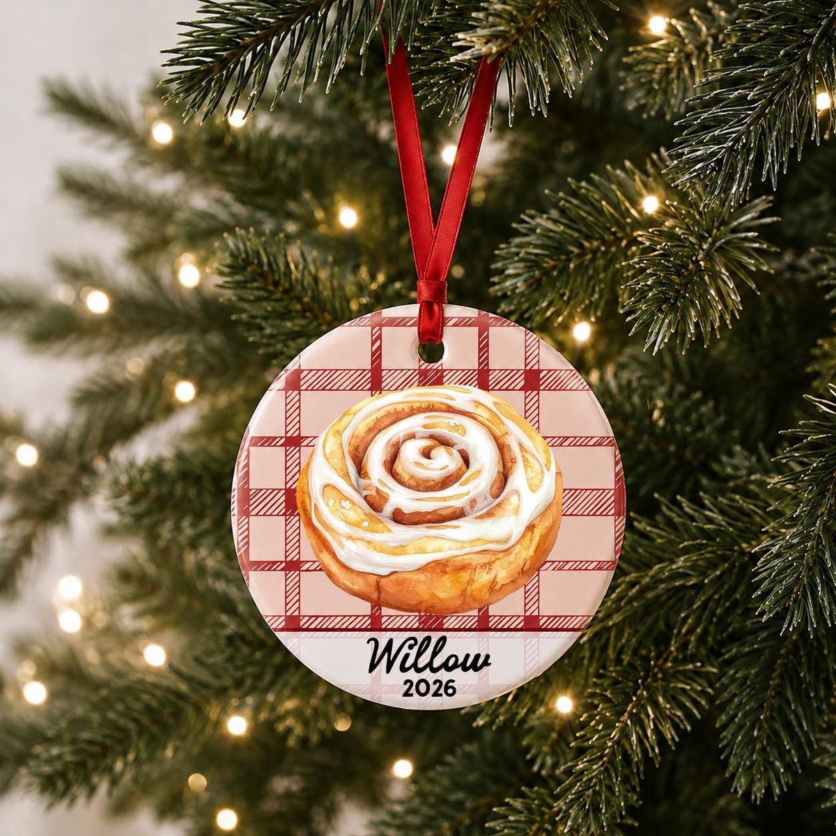 Personalized cinnamon roll ceramic ornament with name and year on red plaid hanging on a lit Christmas tree