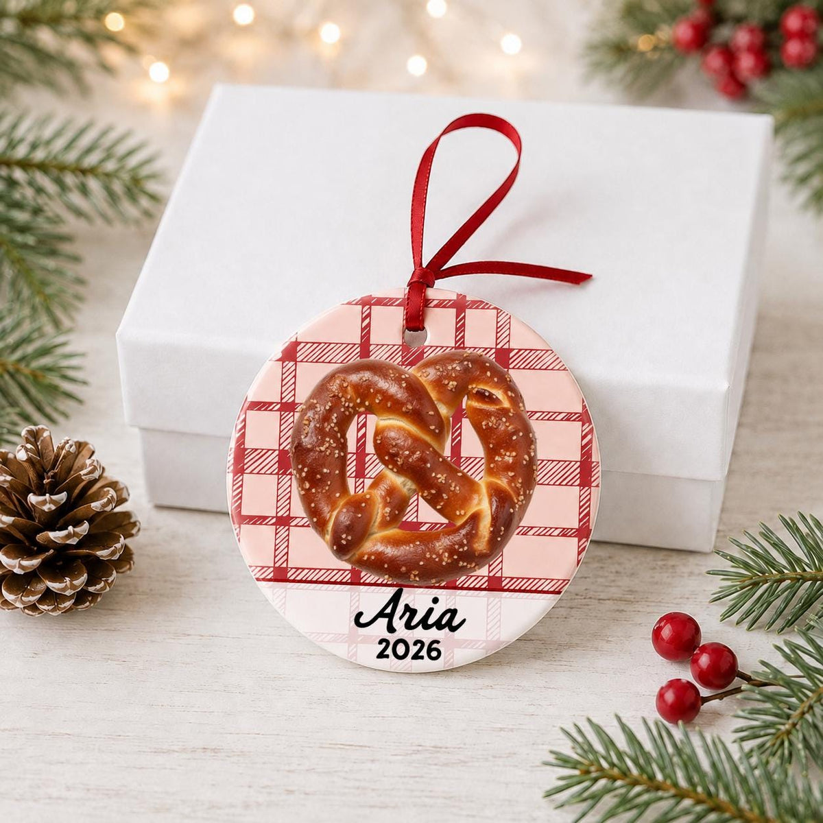 Personalized soft pretzel ceramic ornament with name and year on red plaid in front of a white gift box with pinecones and red berries