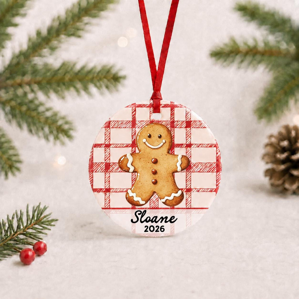 Personalized gingerbread man ceramic ornament with name and year on red plaid with red ribbon on a white surface with pine branches and pinecones
