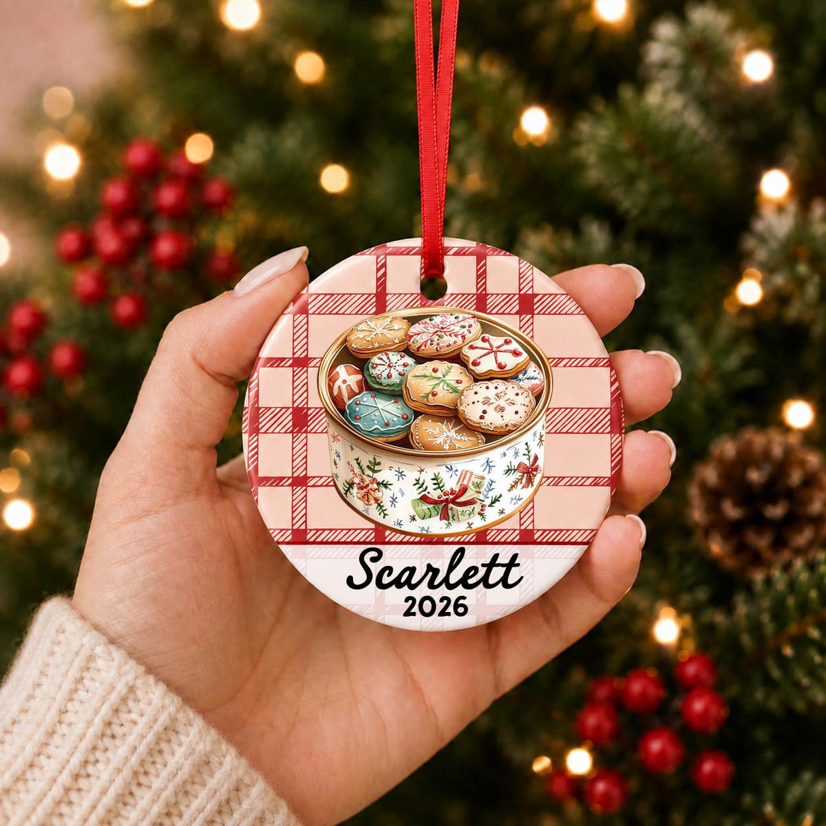 Hand holding the personalized Christmas cookie tin ceramic ornament with name and year on red plaid in front of a Christmas tree