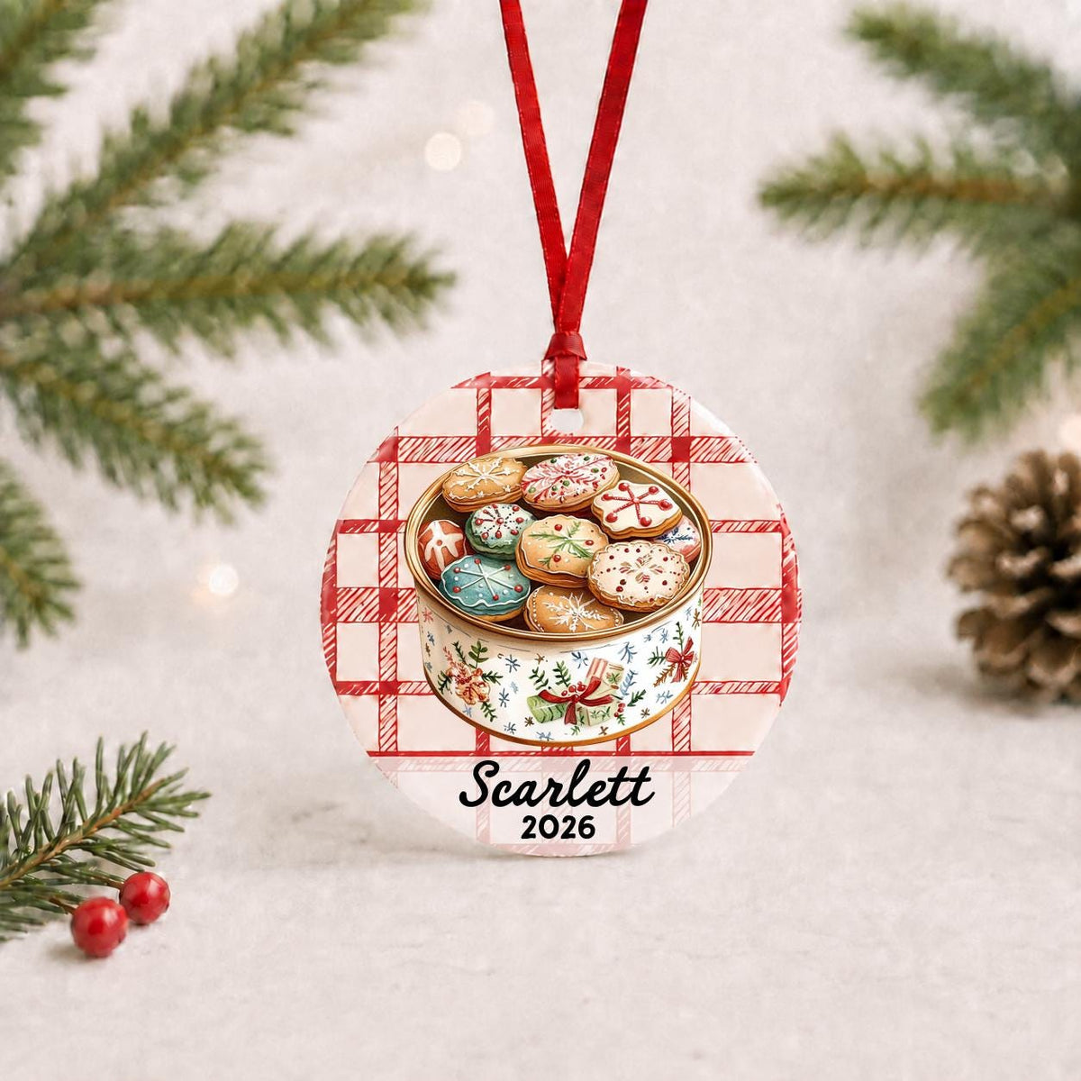 Personalized Christmas cookie tin ceramic ornament with name and year on red plaid with red ribbon on a white surface with pine branches and pinecones