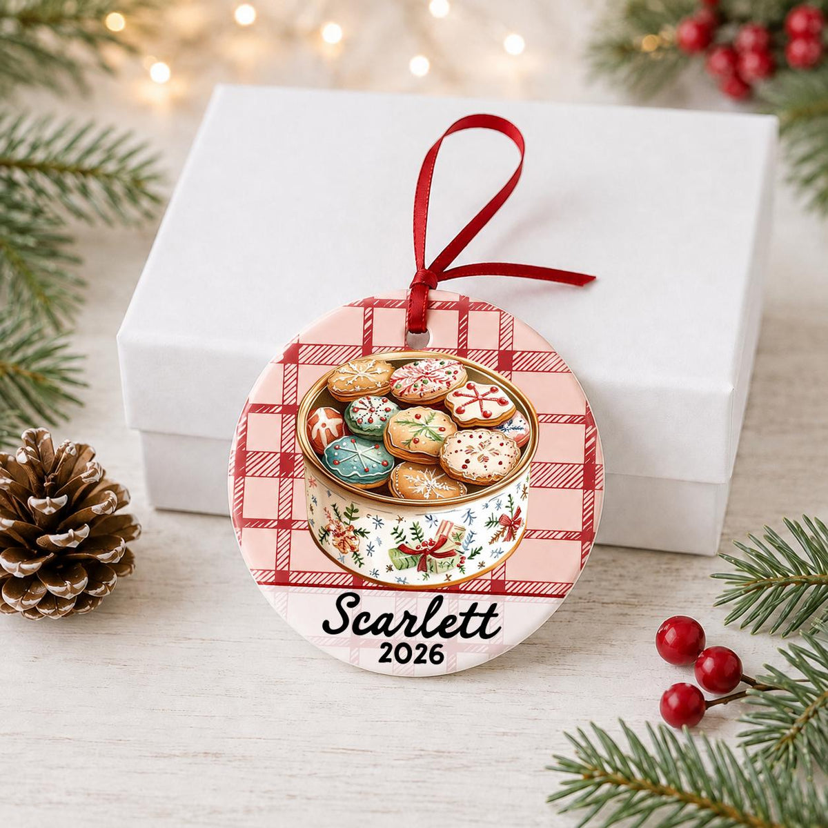 Personalized Christmas cookie tin ceramic ornament with name and year on red plaid in front of a white gift box with pinecones and red berries