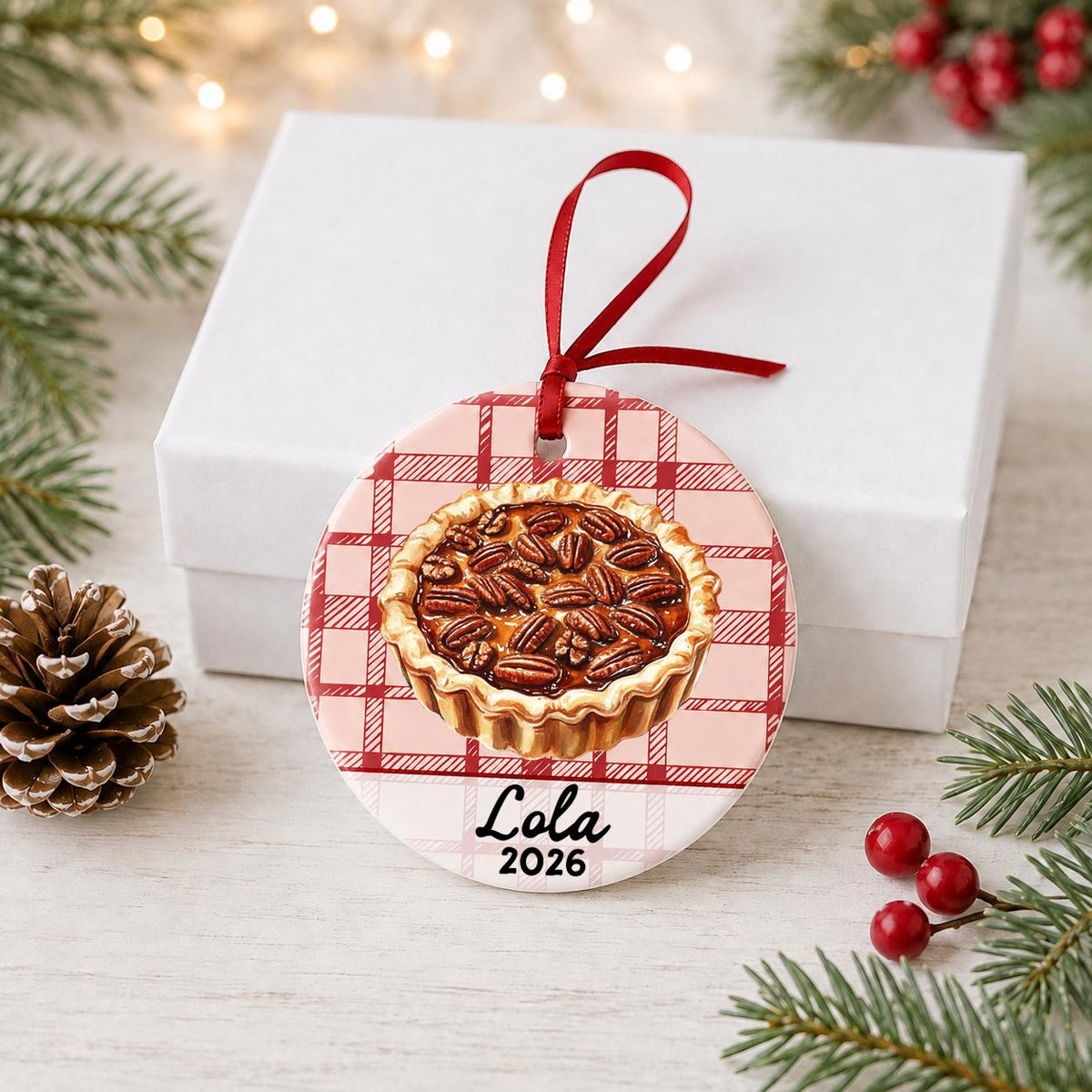 Personalized pecan pie ceramic ornament with name and year on red plaid in front of a white gift box with pinecones and red berries