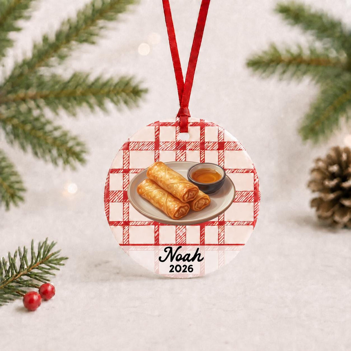 Personalized egg roll ceramic ornament with name and year on red plaid with red ribbon on a white surface with pine branches and pinecones