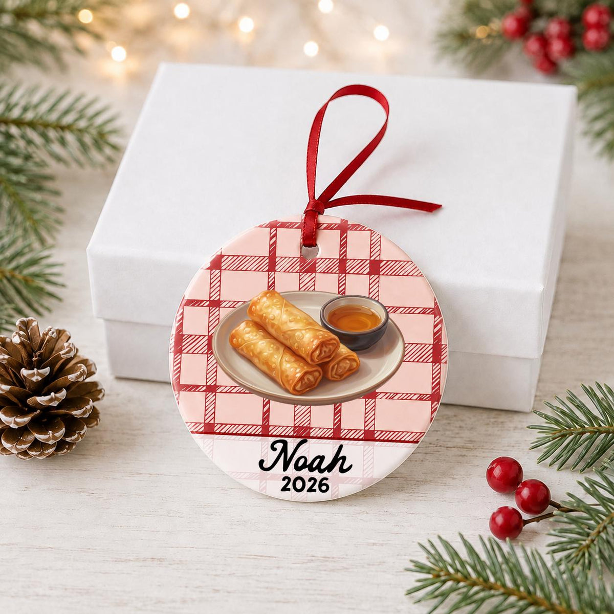 Personalized egg roll ceramic ornament with name and year on red plaid in front of a white gift box with pinecones and red berries