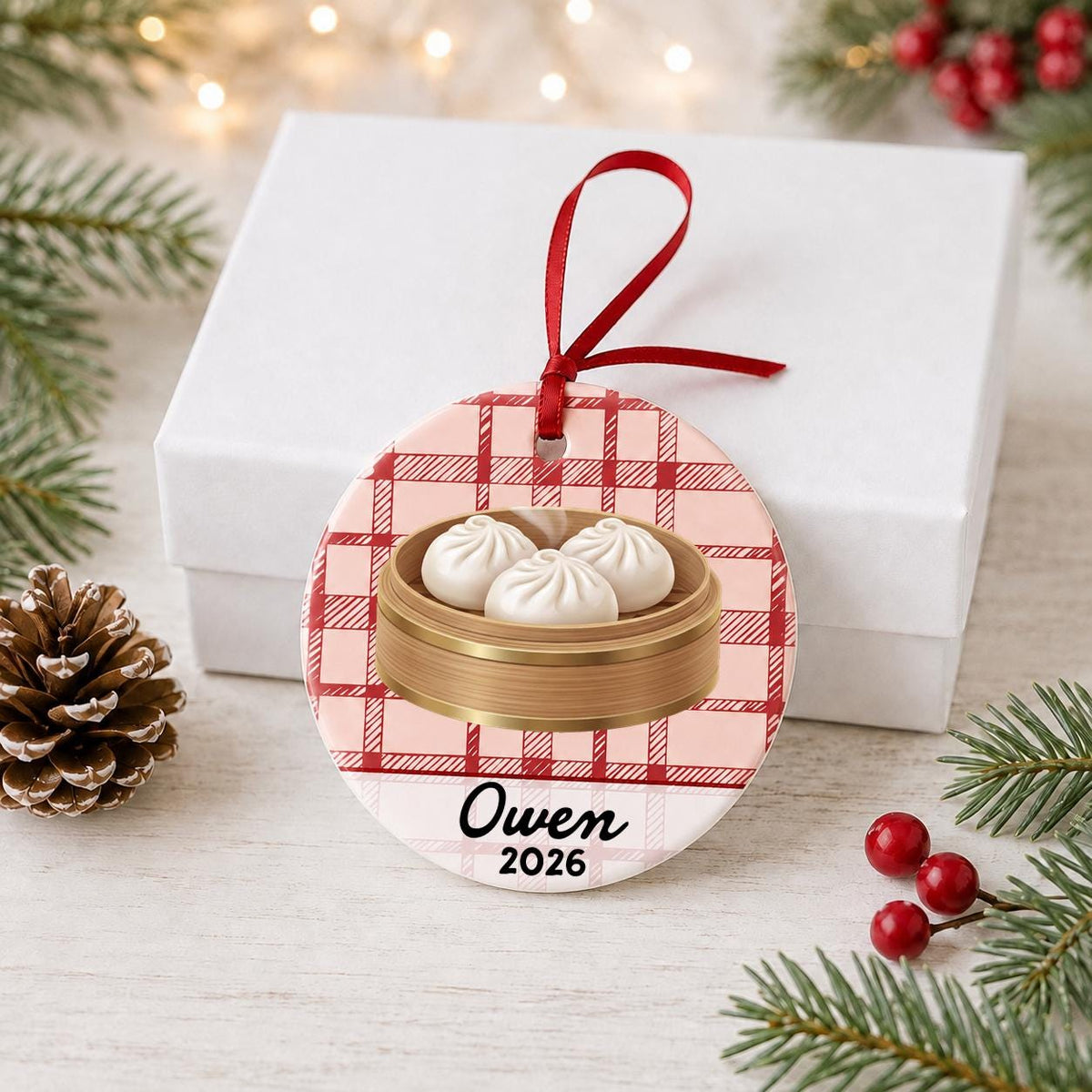 Personalized soup dumpling ceramic ornament with name and year on red plaid in front of a white gift box with pinecones and red berries