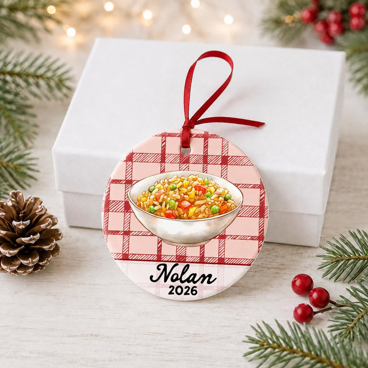 Personalized fried rice ceramic ornament with name and year on red plaid in front of a white gift box with pinecones and red berries
