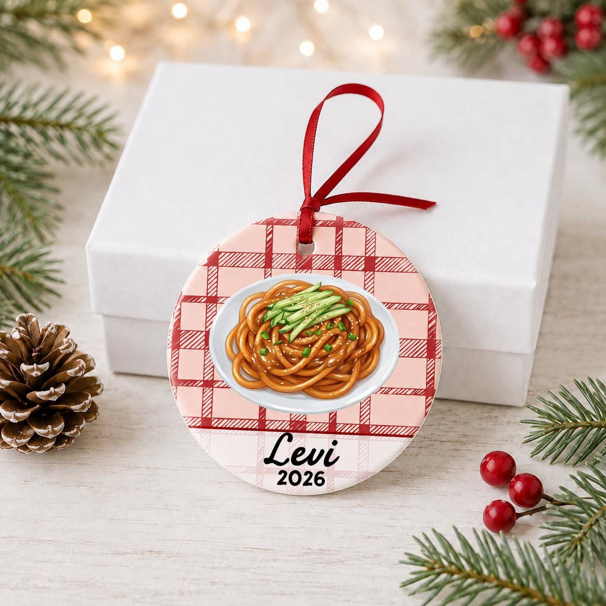 Personalized sesame noodles ceramic ornament with name and year on red plaid in front of a white gift box with pinecones and red berries