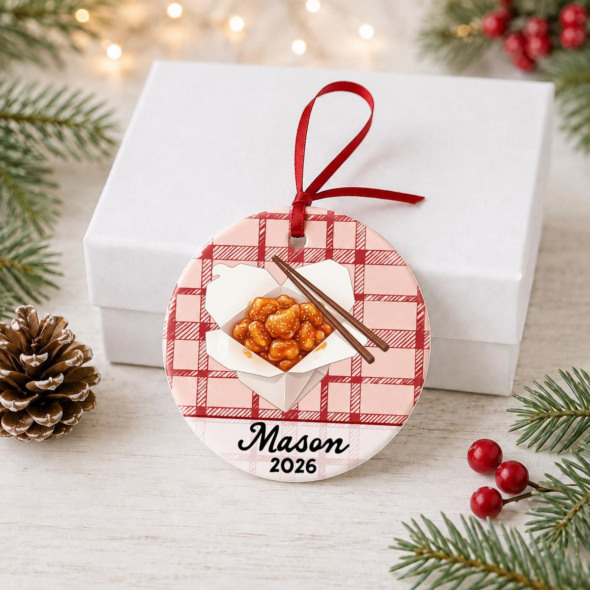 Personalized sesame chicken ceramic ornament with name and year on red plaid in front of a white gift box with pinecones and red berries