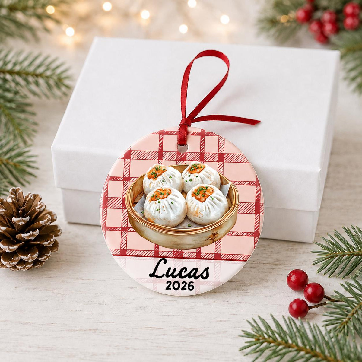 Personalized shumai ceramic ornament with name and year on red plaid in front of a white gift box with pinecones and red berries