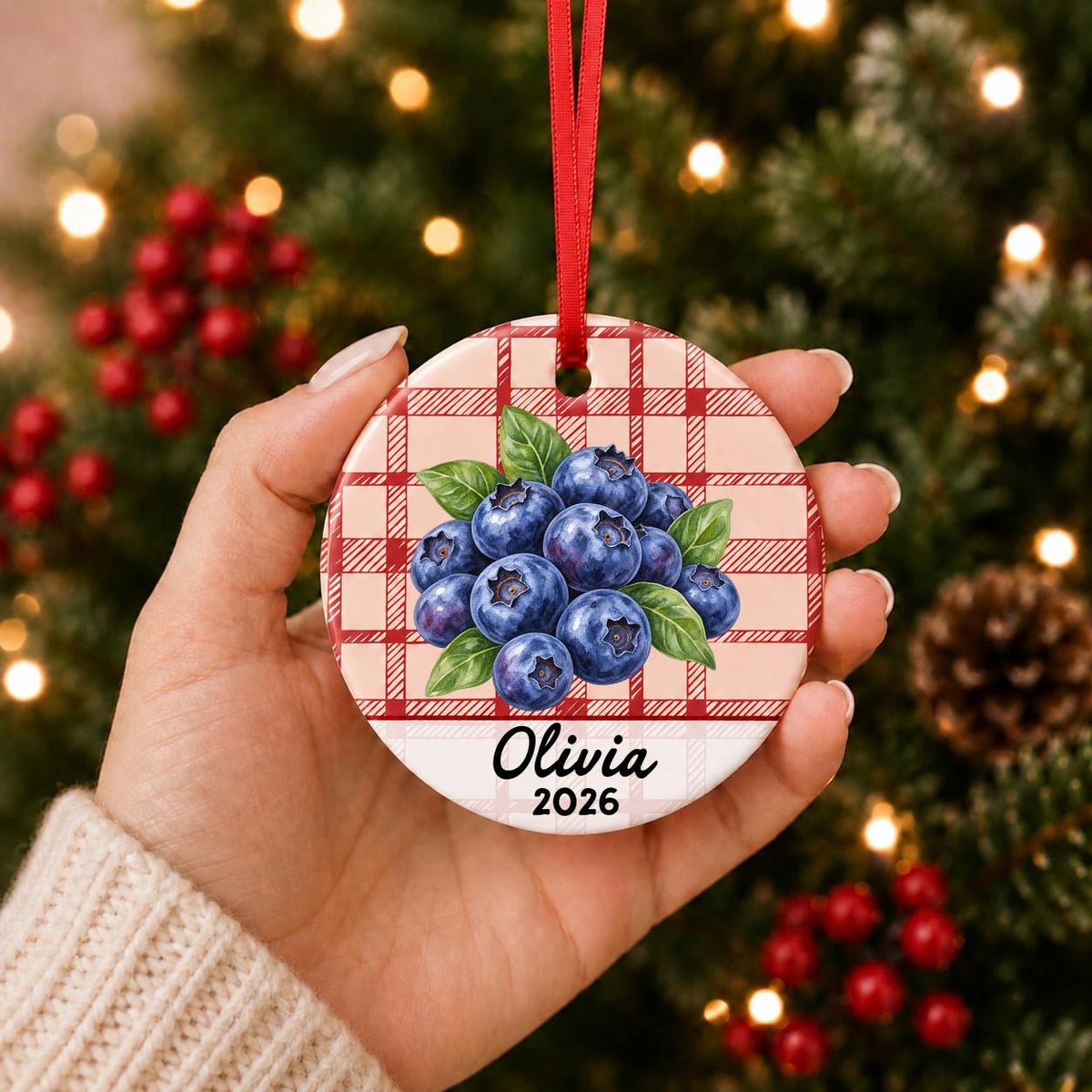 Hand holding the personalized blueberries ceramic ornament with name and year on red plaid in front of a Christmas tree