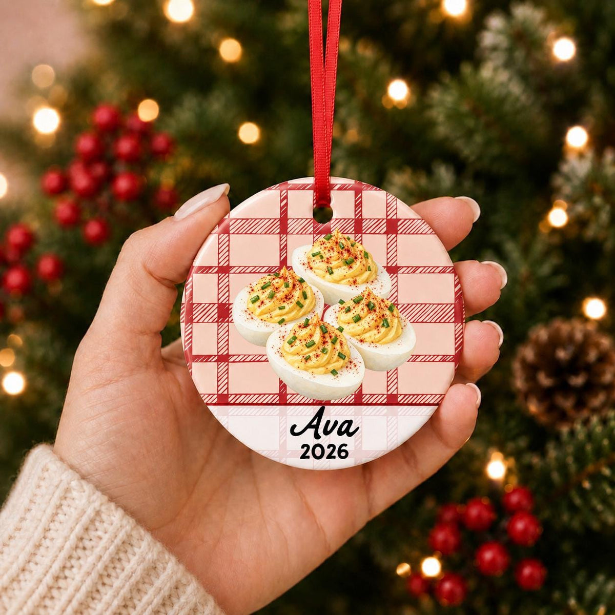 Hand holding the personalized deviled eggs ceramic ornament with name and year on red plaid in front of a Christmas tree