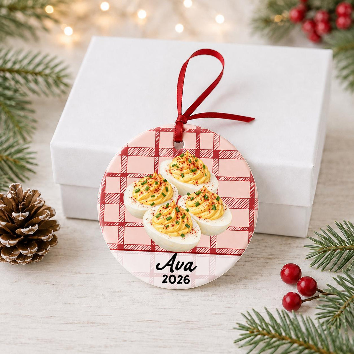 Personalized deviled eggs ceramic ornament with name and year on red plaid in front of a white gift box with pinecones and red berries