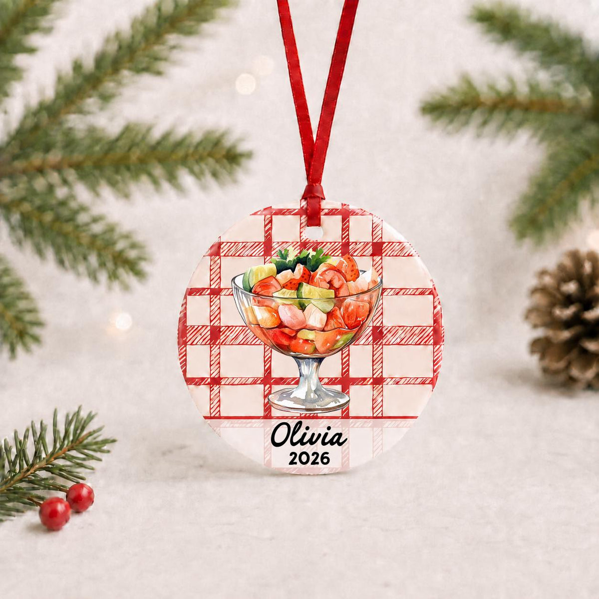 Personalized shrimp cocktail ceramic ornament with name and year on red plaid with red ribbon on a white surface with pine branches and pinecones