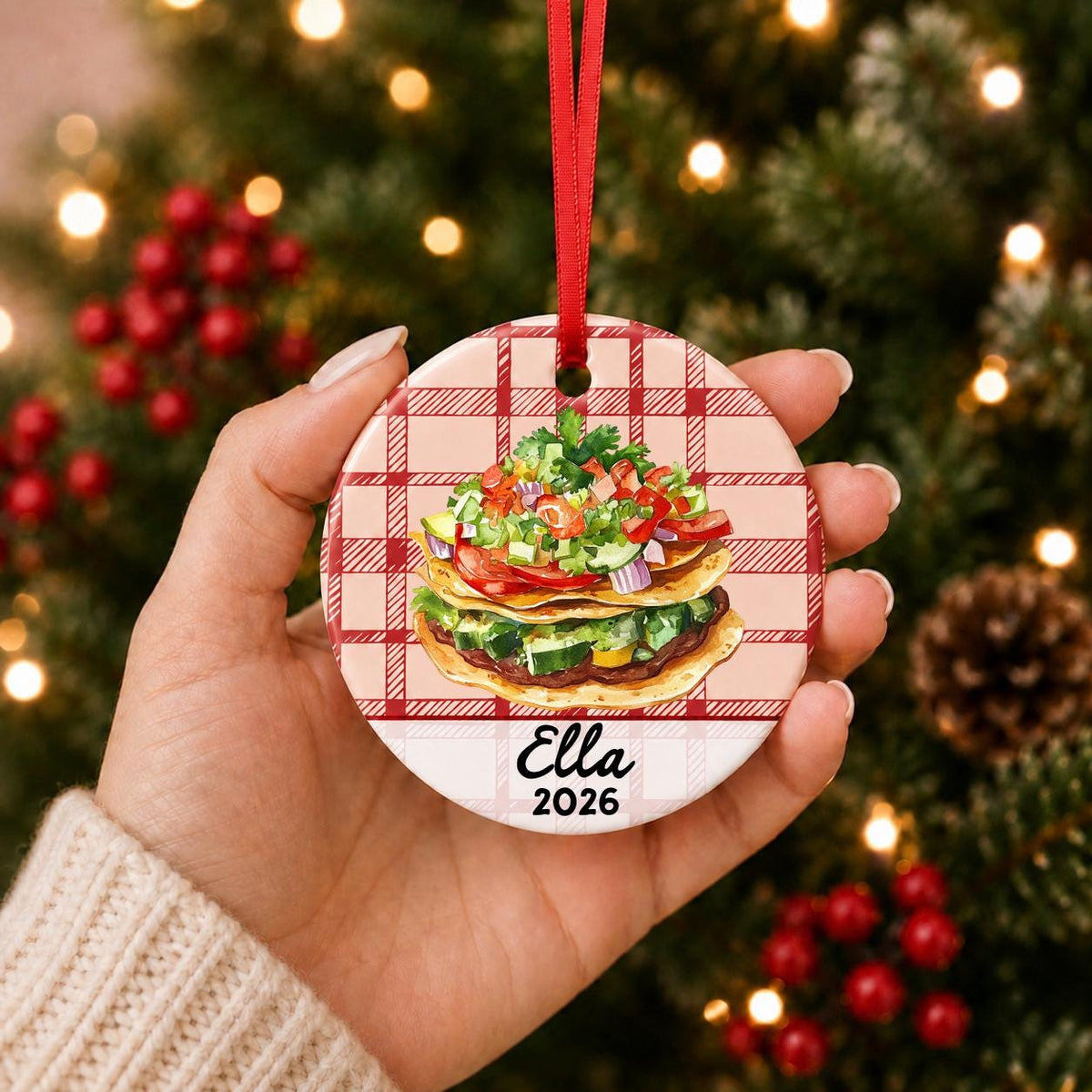 Hand holding the personalized tostada ceramic ornament with name and year on red plaid in front of a Christmas tree