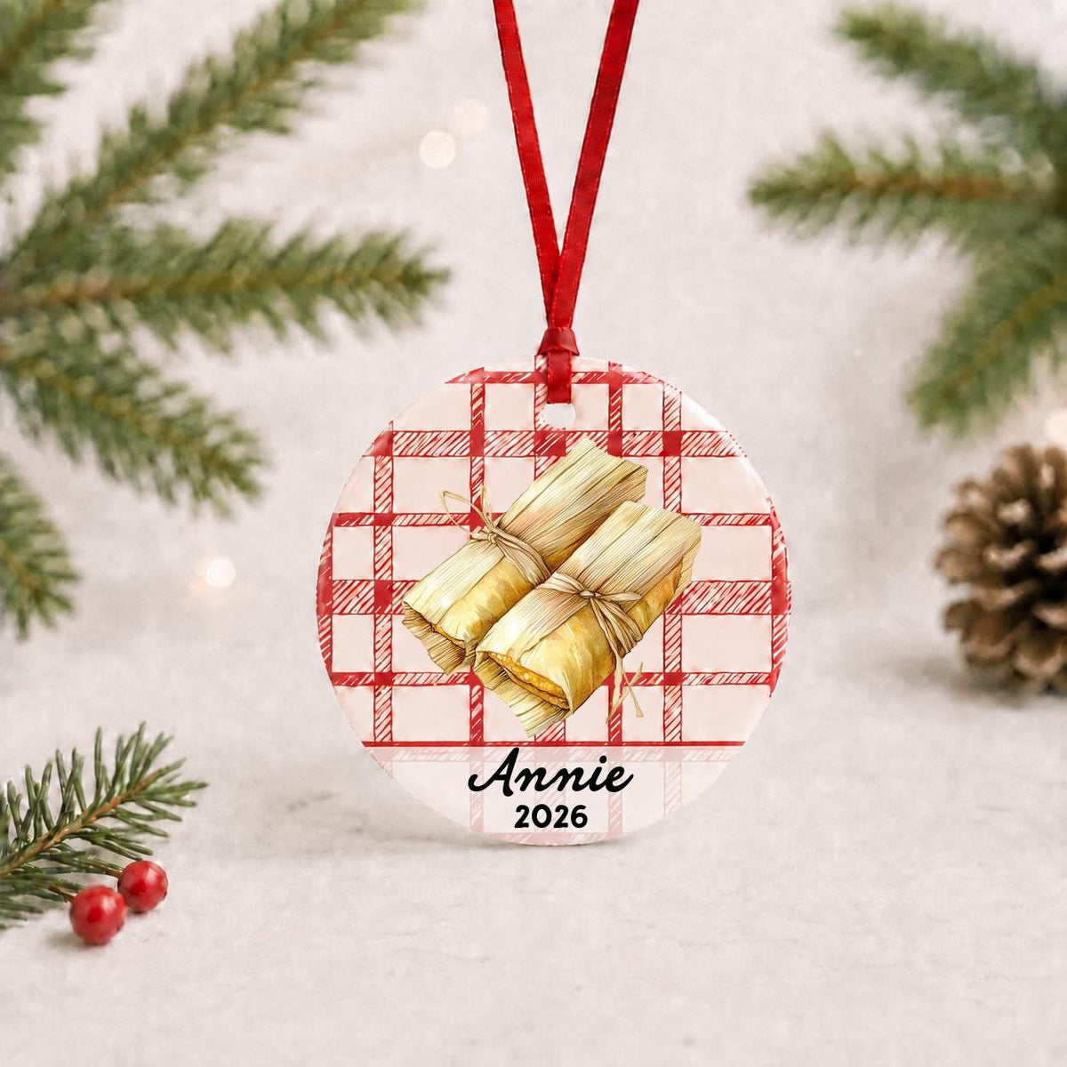 Personalized tamale ceramic ornament with name and year on red plaid with red ribbon on a white surface with pine branches and pinecones