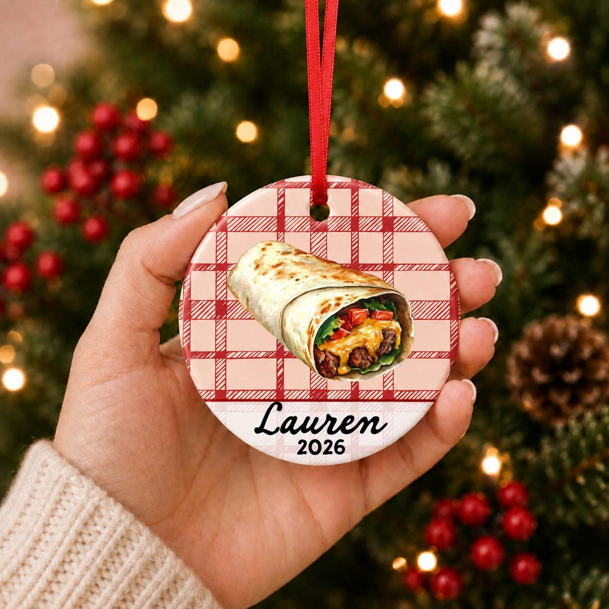 Hand holding the personalized burrito ceramic ornament with name and year on red plaid in front of a Christmas tree