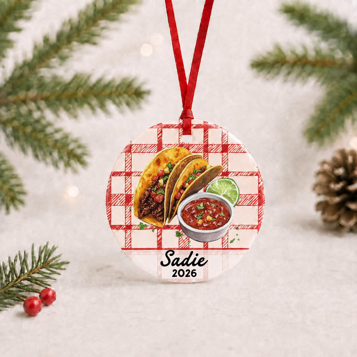 Personalized taco ceramic ornament with name and year on red plaid with red ribbon on a white surface with pine branches and pinecones