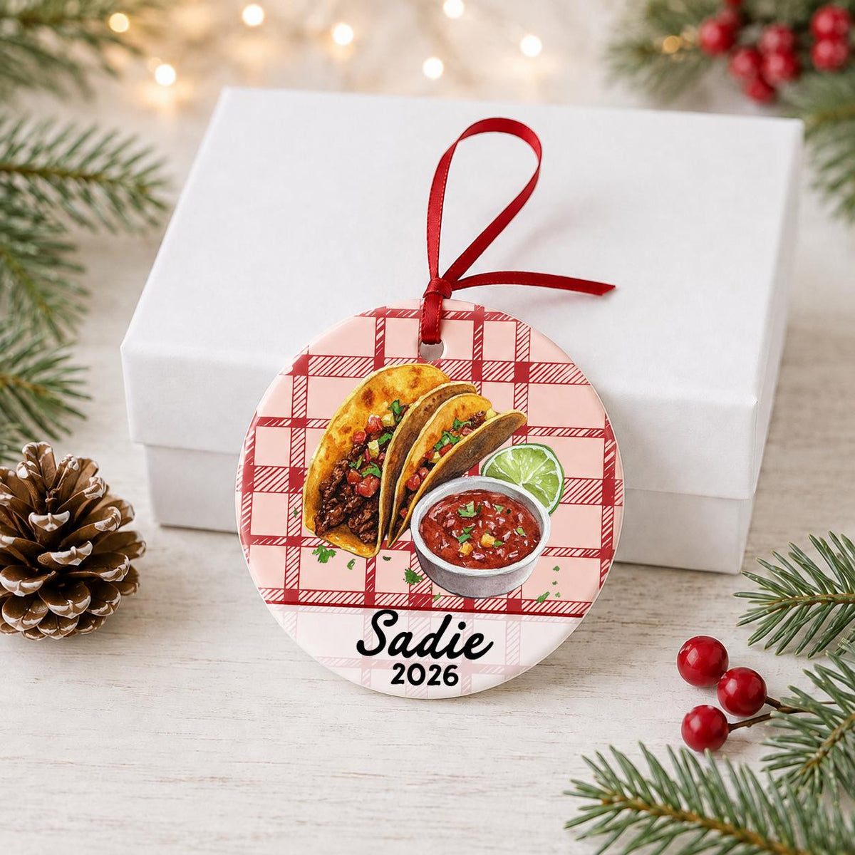 Personalized taco ceramic ornament with name and year on red plaid in front of a white gift box with pinecones and red berries
