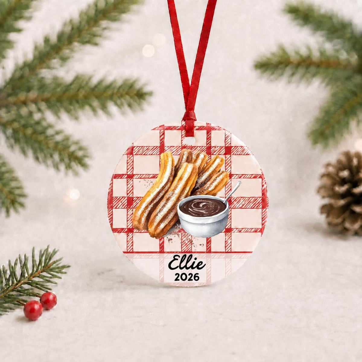 Personalized churro ceramic ornament with name and year on red plaid with red ribbon on a white surface with pine branches and pinecones