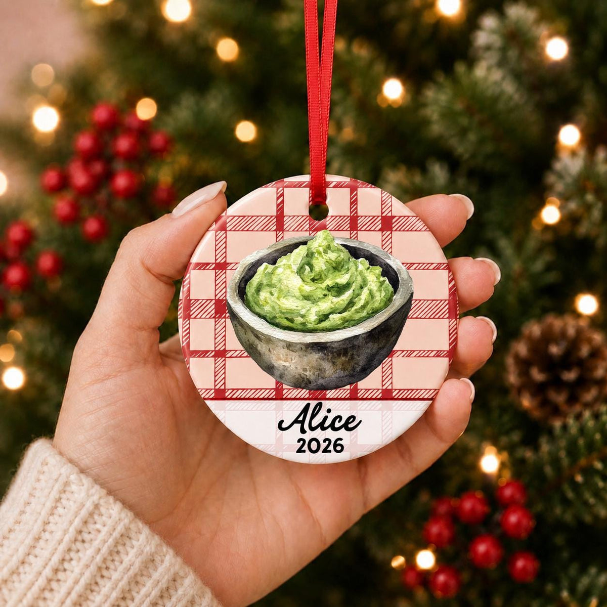 Hand holding the personalized guacamole ceramic ornament with name and year on red plaid in front of a Christmas tree