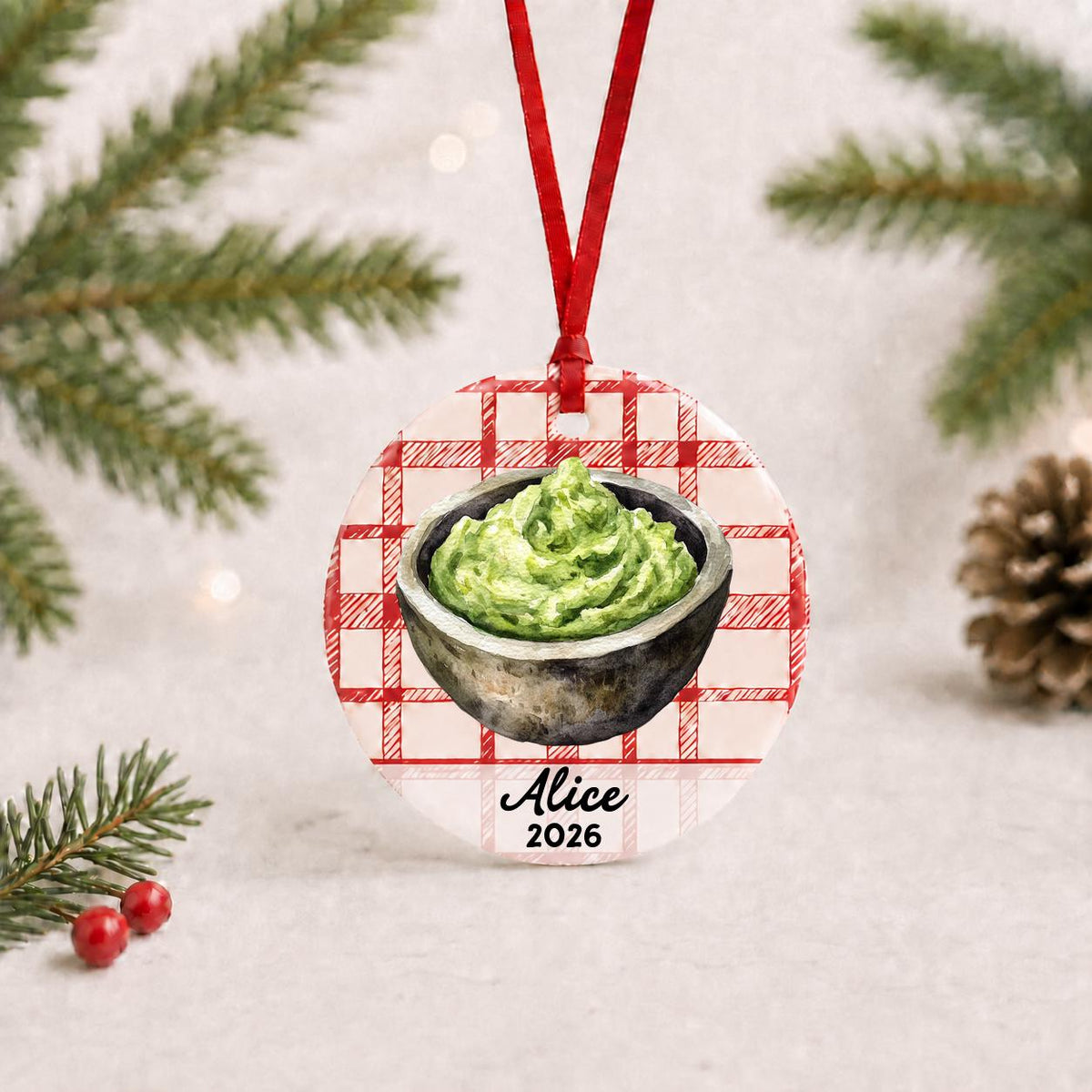 Personalized guacamole ceramic ornament with name and year on red plaid with red ribbon on a white surface with pine branches and pinecones