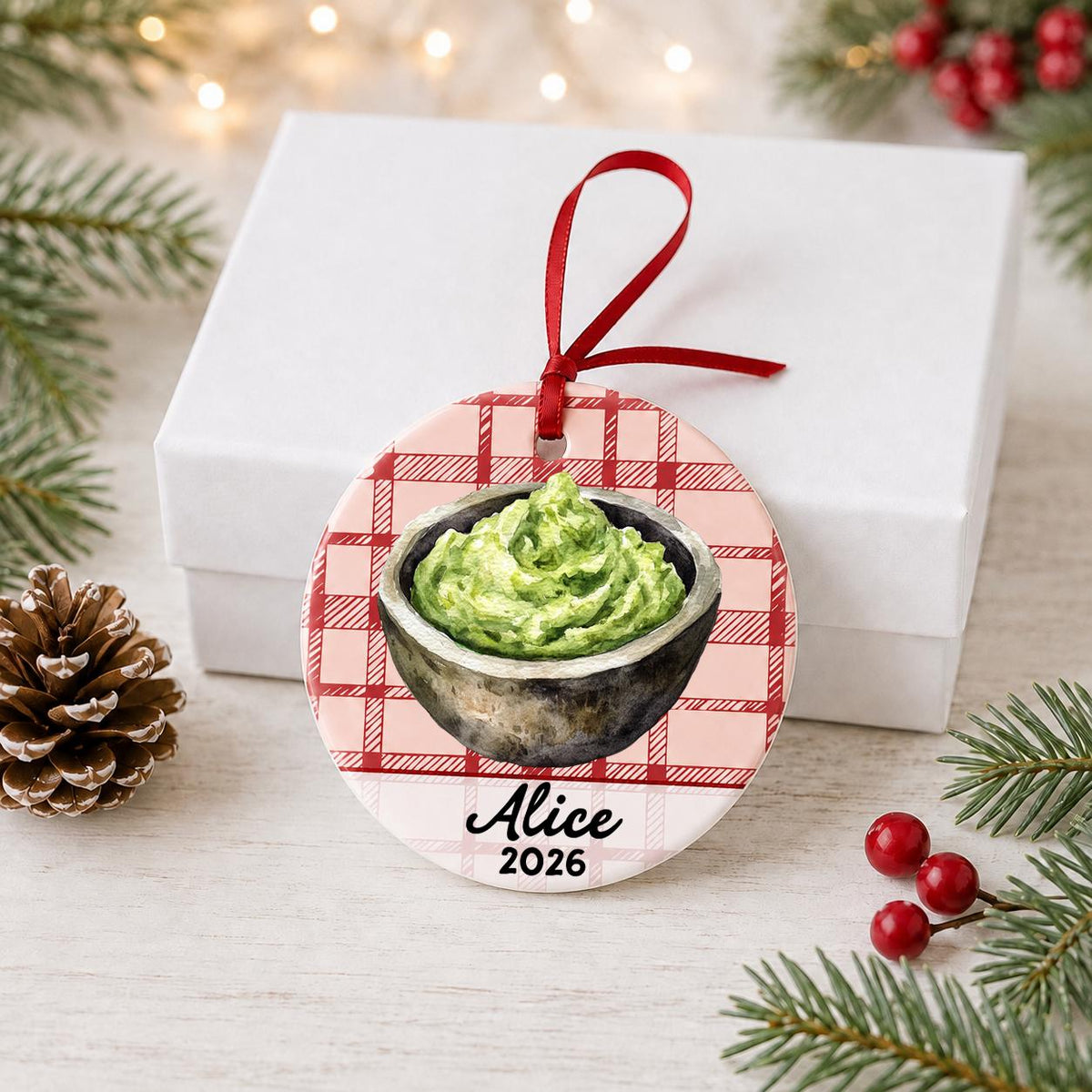 Personalized guacamole ceramic ornament with name and year on red plaid in front of a white gift box with pinecones and red berries