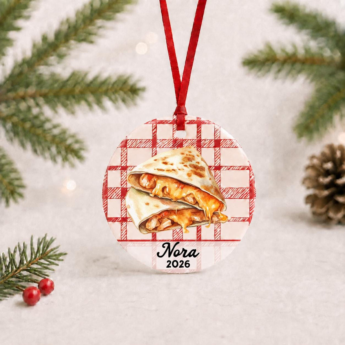 Personalized quesadilla ceramic ornament with name and year on red plaid with red ribbon on a white surface with pine branches and pinecones