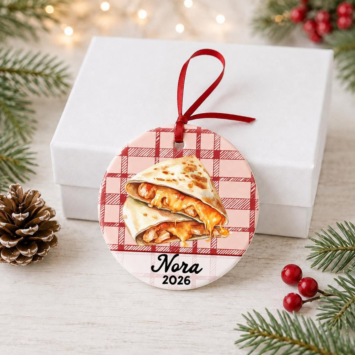 Personalized quesadilla ceramic ornament with name and year on red plaid in front of a white gift box with pinecones and red berries