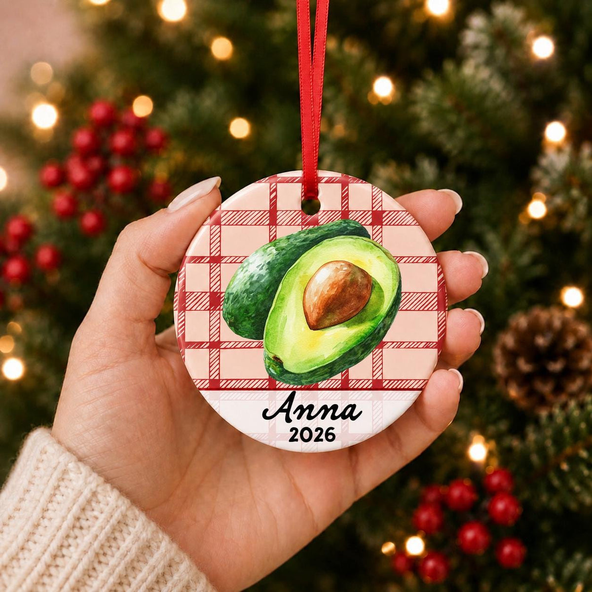 Hand holding the personalized avocado ceramic ornament with name and year on red plaid in front of a Christmas tree
