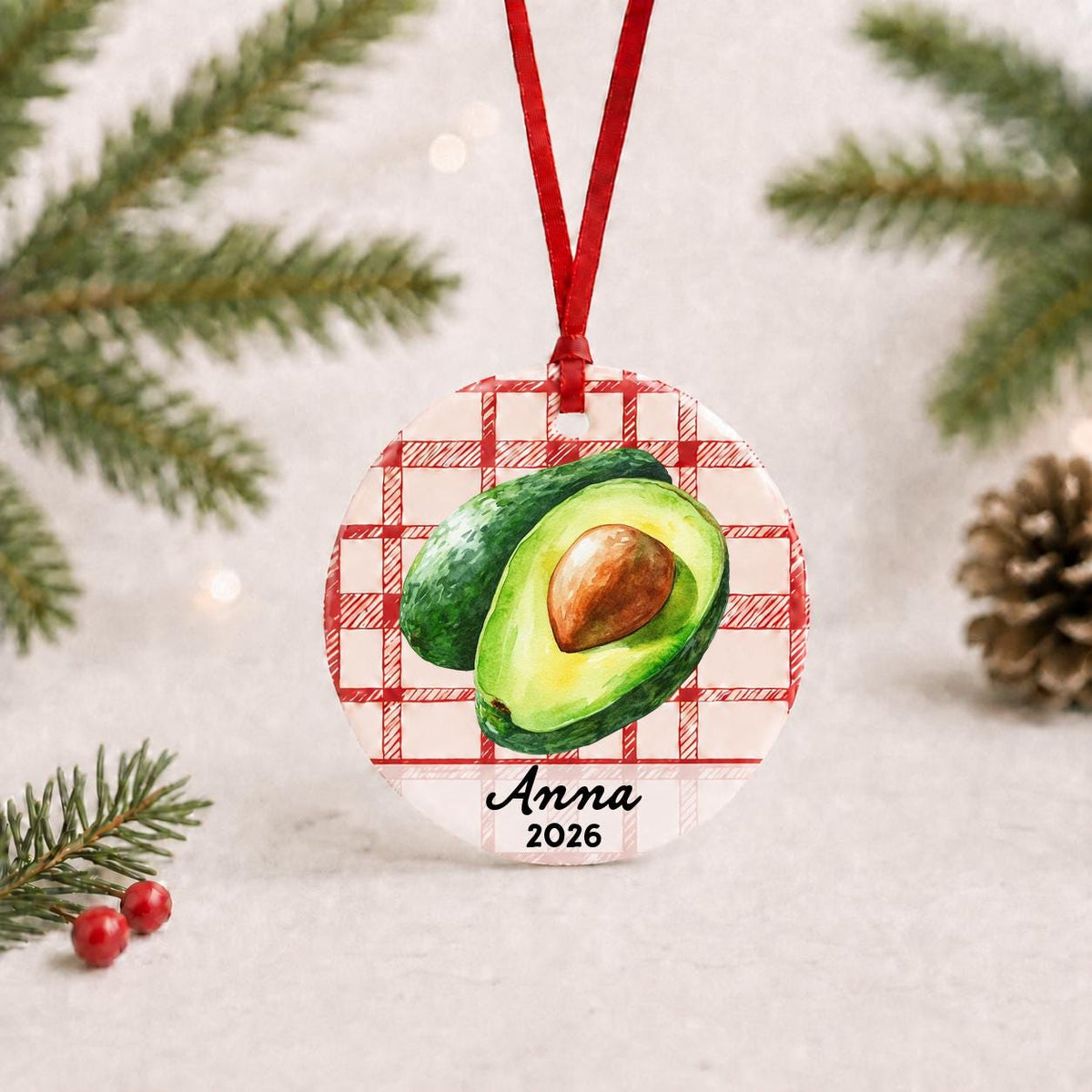 Personalized avocado ceramic ornament with name and year on red plaid with red ribbon on a white surface with pine branches and pinecones