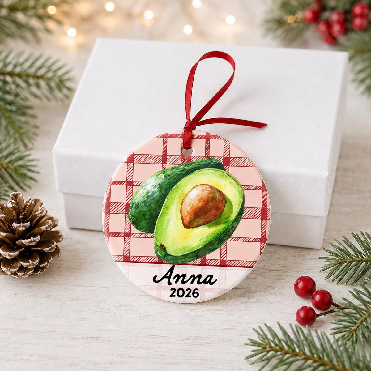 Personalized avocado ceramic ornament with name and year on red plaid in front of a white gift box with pinecones and red berries