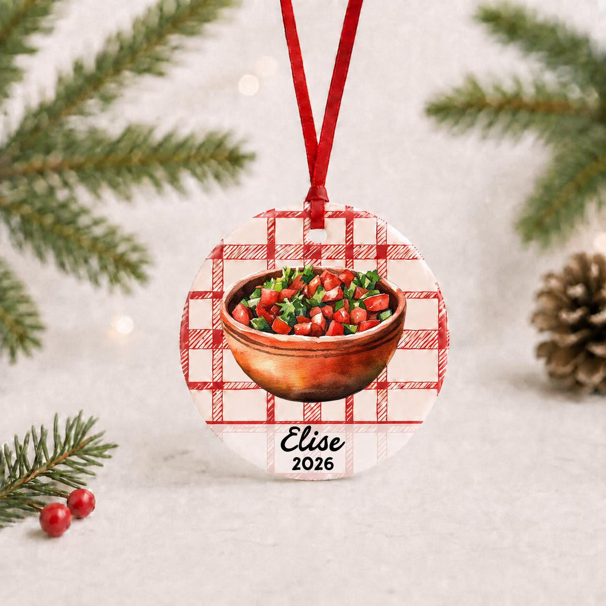 Personalized pico de gallo ceramic ornament with name and year on red plaid with red ribbon on a white surface with pine branches and pinecones
