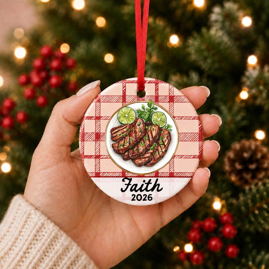 Hand holding the personalized carne asada ceramic ornament with name and year on red plaid in front of a Christmas tree