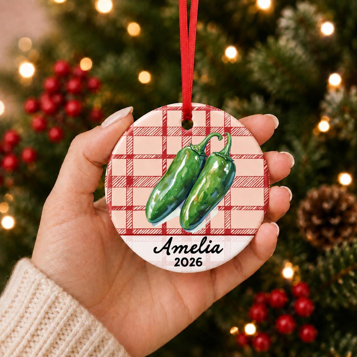 Hand holding the personalized jalapeño ceramic ornament with name and year on red plaid in front of a Christmas tree