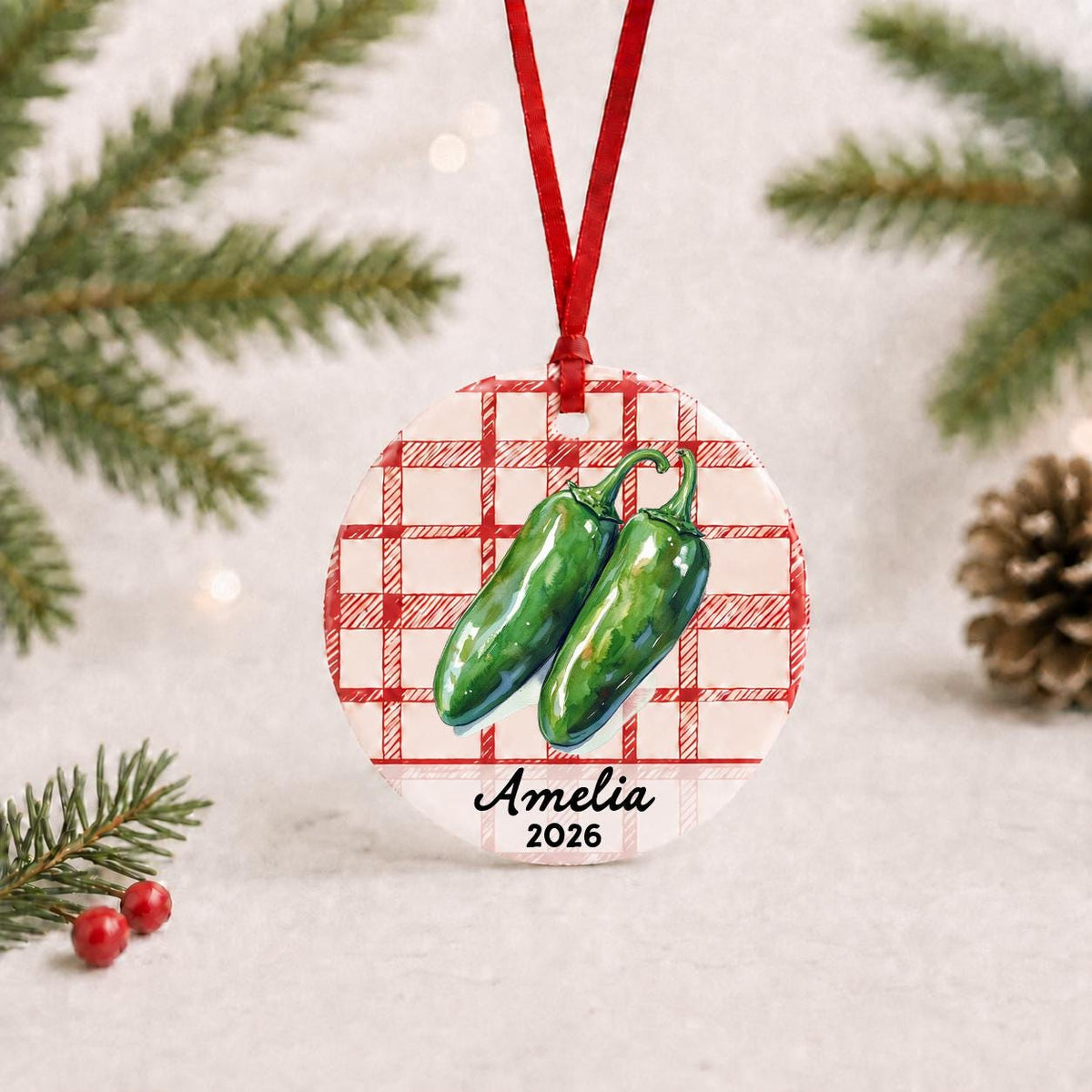Personalized jalapeño ceramic ornament with name and year on red plaid with red ribbon on a white surface with pine branches and pinecones