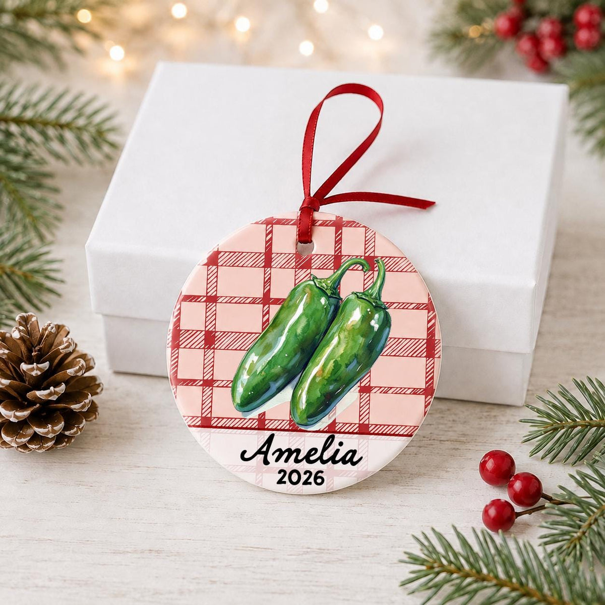 Personalized jalapeño ceramic ornament with name and year on red plaid in front of a white gift box with pinecones and red berries