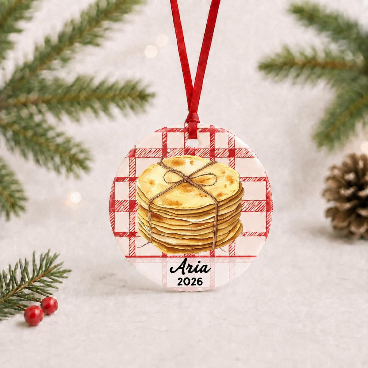Personalized tortilla ceramic ornament with name and year on red plaid with red ribbon on a white surface with pine branches and pinecones