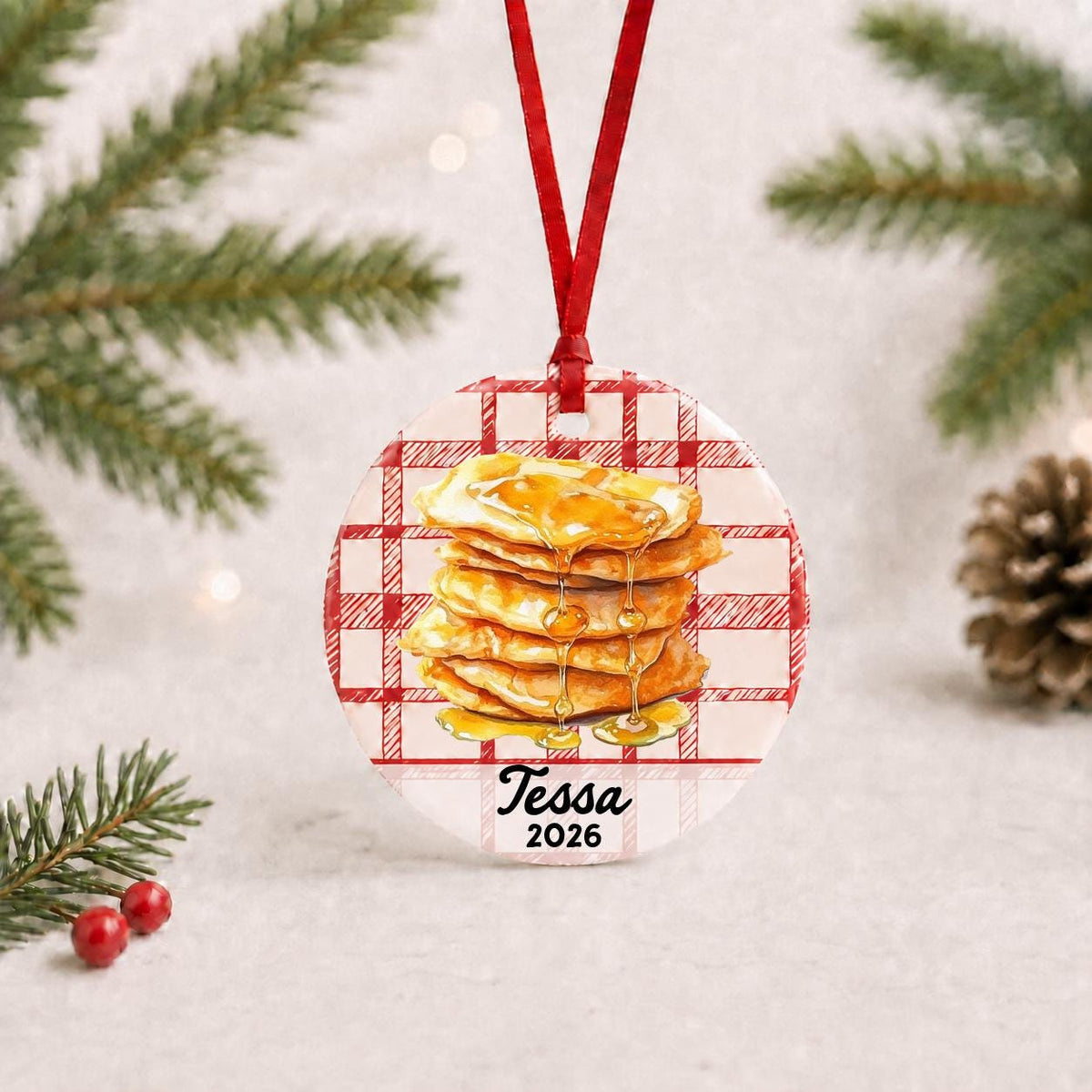 Personalized buñuelos ceramic ornament with name and year on red plaid with red ribbon on a white surface with pine branches and pinecones
