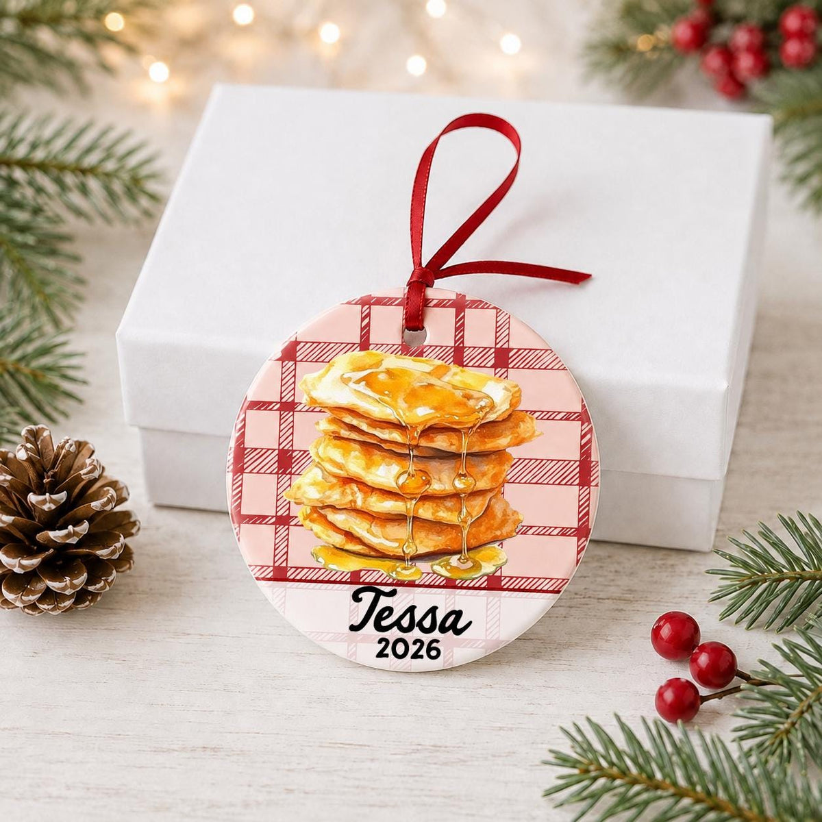 Personalized buñuelos ceramic ornament with name and year on red plaid in front of a white gift box with pinecones and red berries