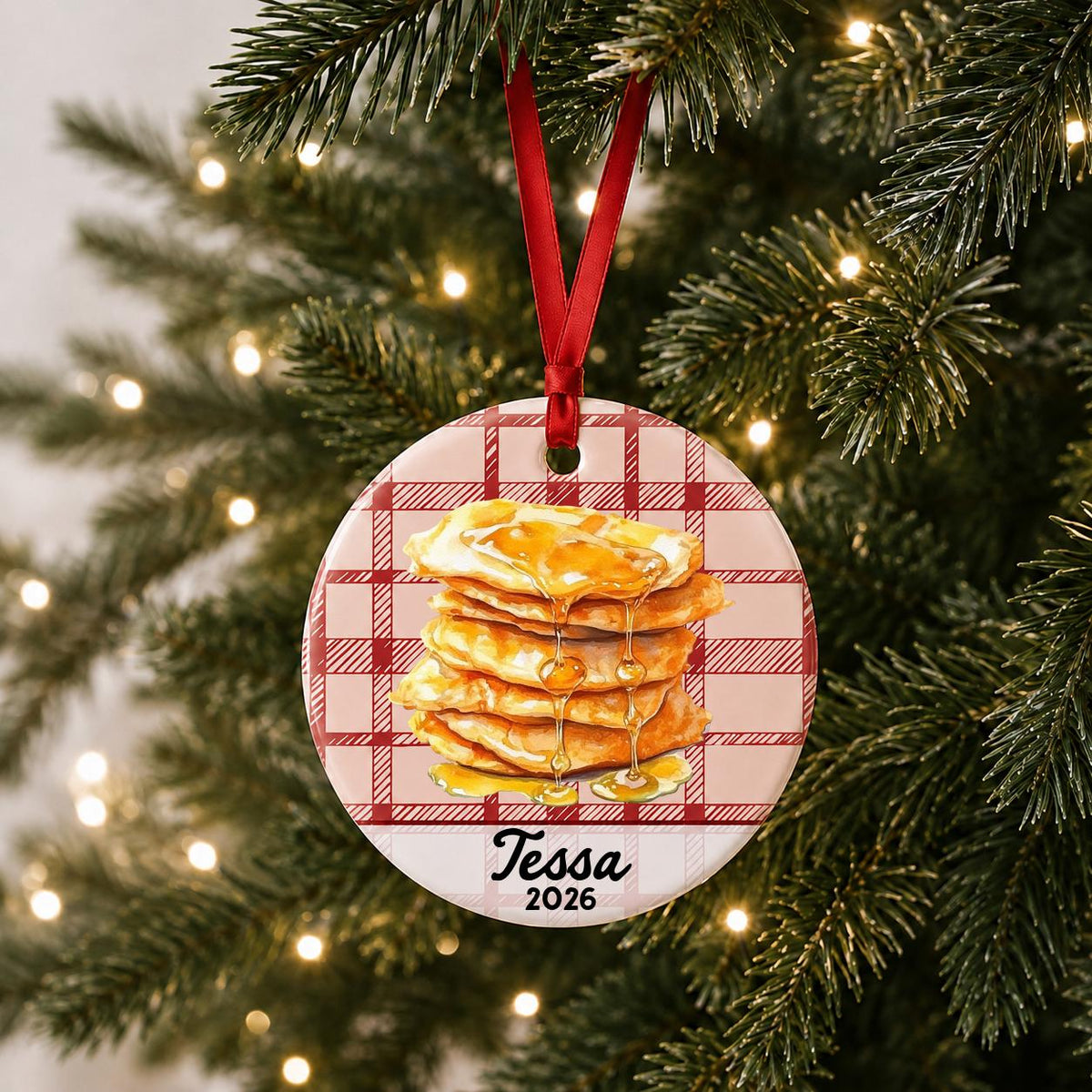 Personalized buñuelos ceramic ornament with name and year on red plaid hanging on a lit Christmas tree
