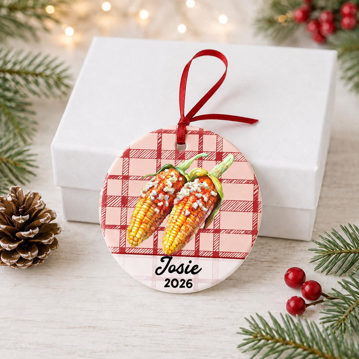 Personalized Mexican street corn ceramic ornament with name and year on red plaid in front of a white gift box with pinecones and red berries