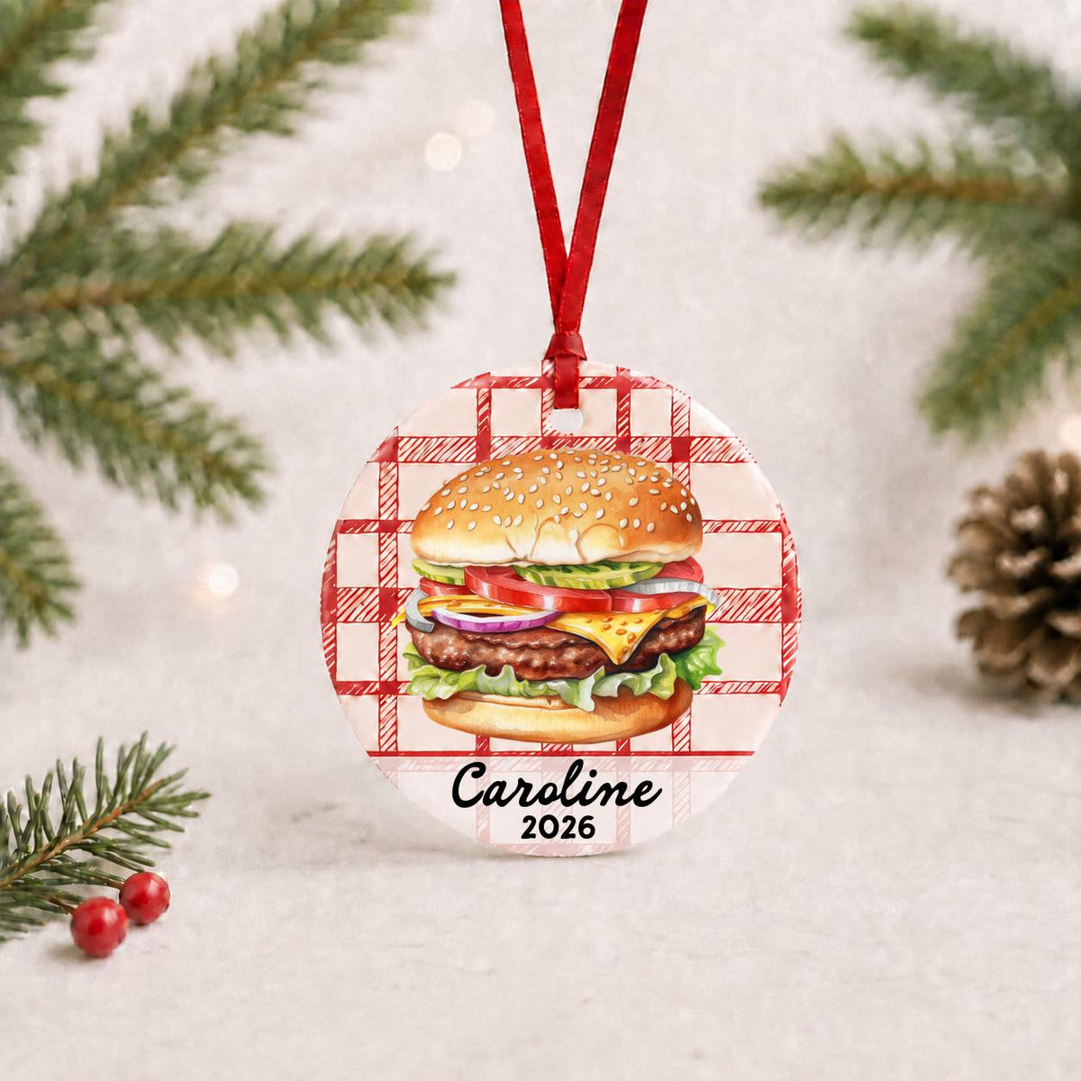 Personalized cheeseburger ceramic ornament with name and year on red plaid with red ribbon on a white surface with pine branches and pinecones