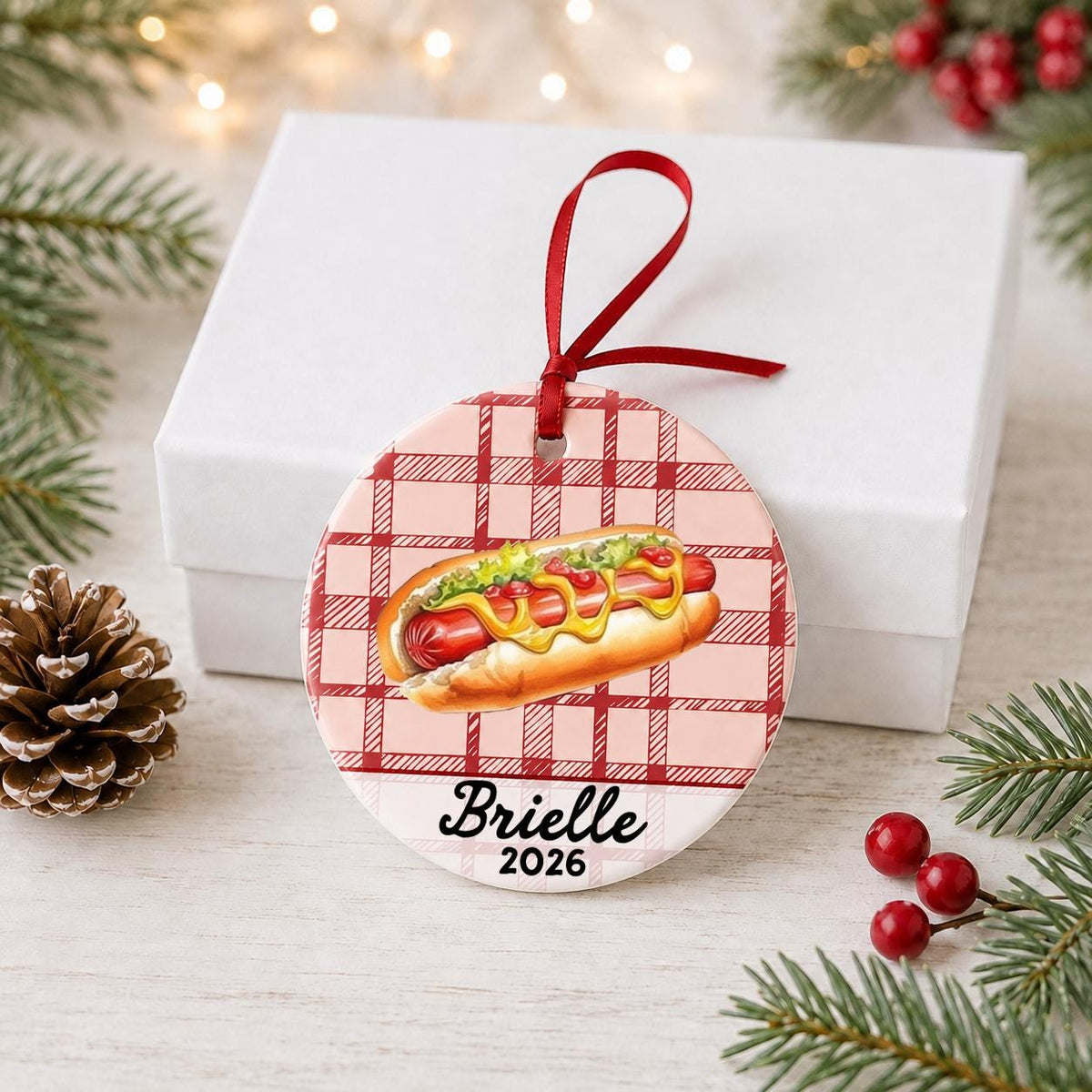 Personalized hot dog ceramic ornament with name and year on red plaid in front of a white gift box with pinecones and red berries