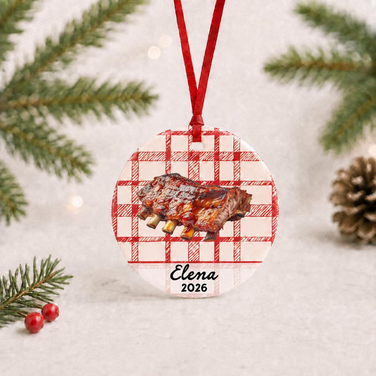 Personalized BBQ ribs ceramic ornament with name and year on red plaid with red ribbon on a white surface with pine branches and pinecones
