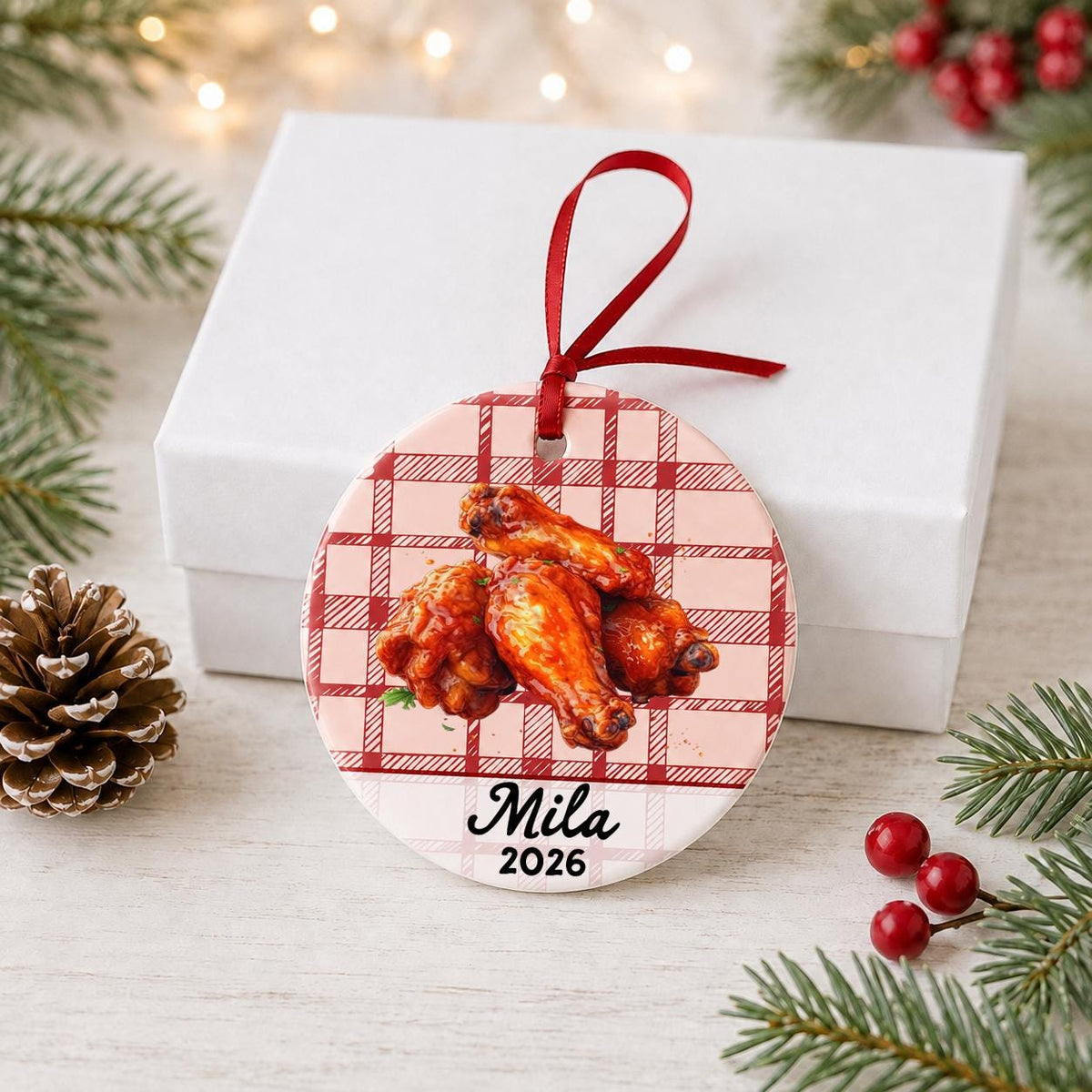 Personalized buffalo wings ceramic ornament with name and year on red plaid in front of a white gift box with pinecones and red berries