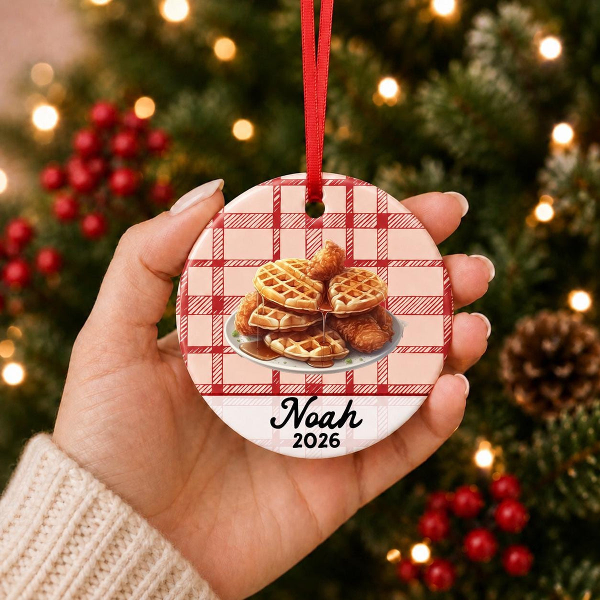 Hand holding the personalized chicken and waffles ceramic ornament with name and year on red plaid in front of a Christmas tree