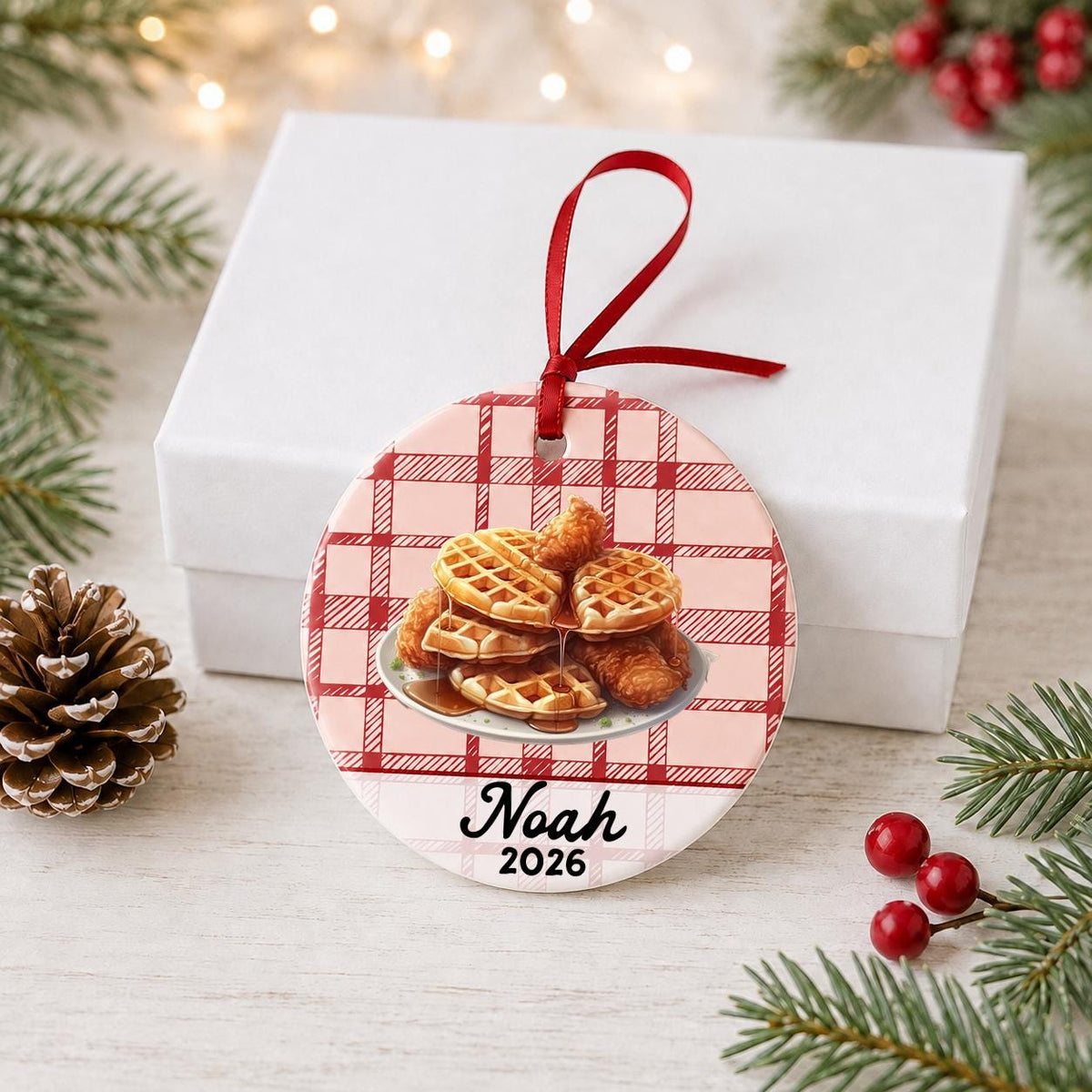 Personalized chicken and waffles ceramic ornament with name and year on red plaid in front of a white gift box with pinecones and red berries